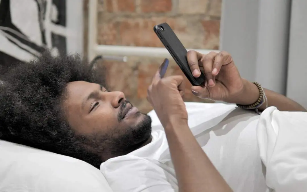 A man with curly hair lies on a bed, holding a smartphone and looking at it.