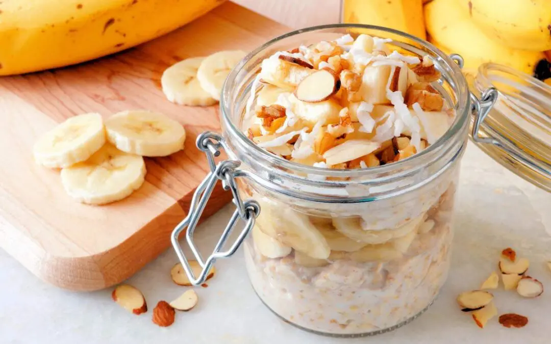 A glass jar filled with layered oatmeal, sliced bananas, and toppings such as almonds and coconut flakes, with a wooden cutting board and whole bananas in the background.
