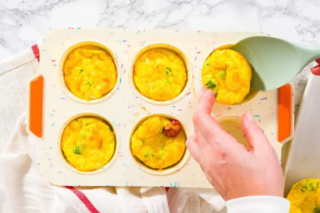 A hand using a spatula to remove baked egg muffins from a colorful muffin tray on a marble countertop.