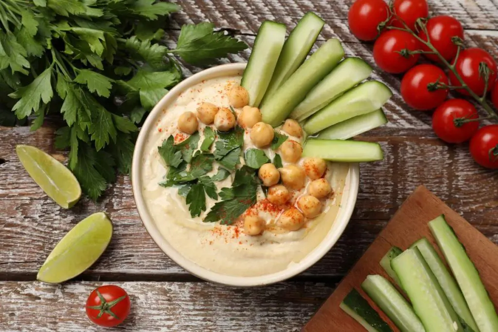A bowl of hummus topped with chickpeas and parsley, surrounded by sliced cucumbers, cherry tomatoes, lime wedges, and fresh herbs on a rustic wooden surface.