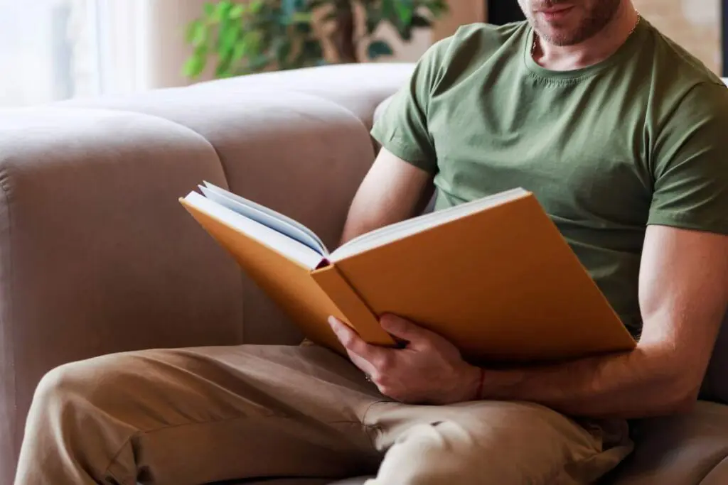 A person sitting on a couch, reading a large book with an orange cover.