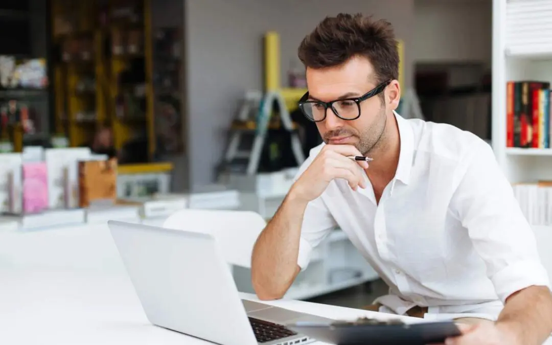 A young man wearing glasses is sitting at a table, looking thoughtfully at a laptop while holding a pen and clipboard.