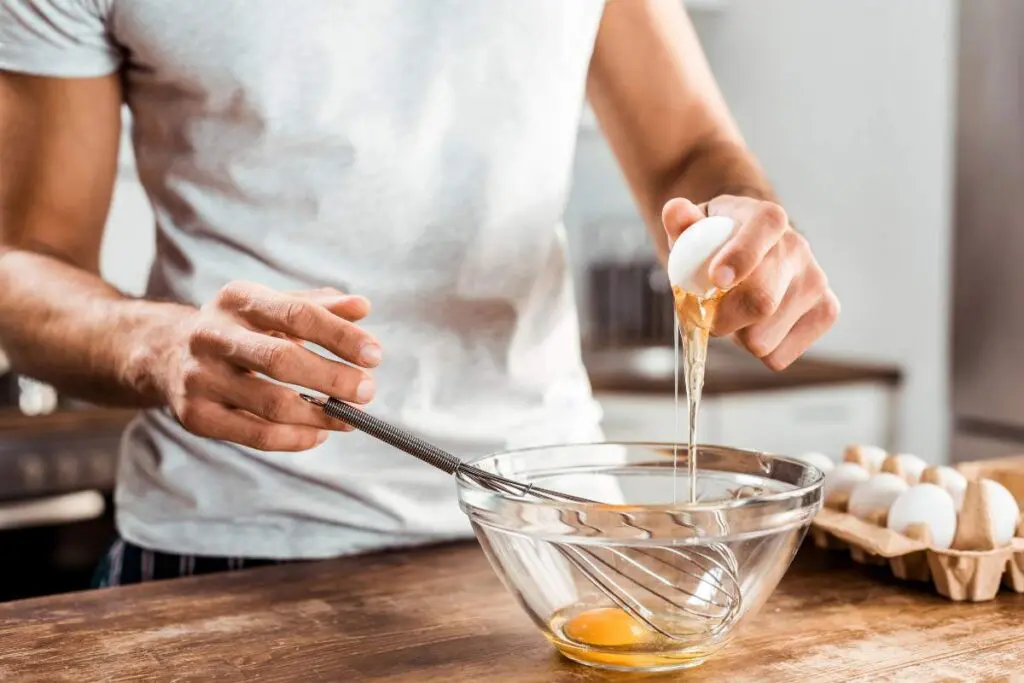 A person cracking an egg into a glass bowl while holding a whisk, with a carton of eggs in the background.