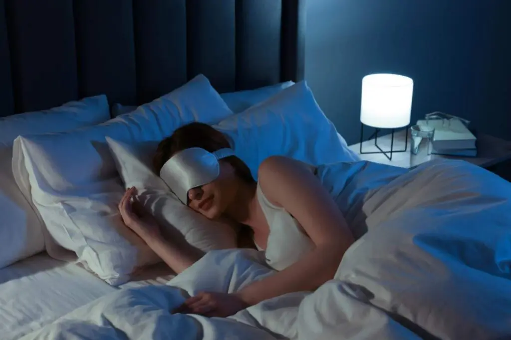 A person sleeping in bed wearing an eye mask, with a soft glow from a lamp beside them.