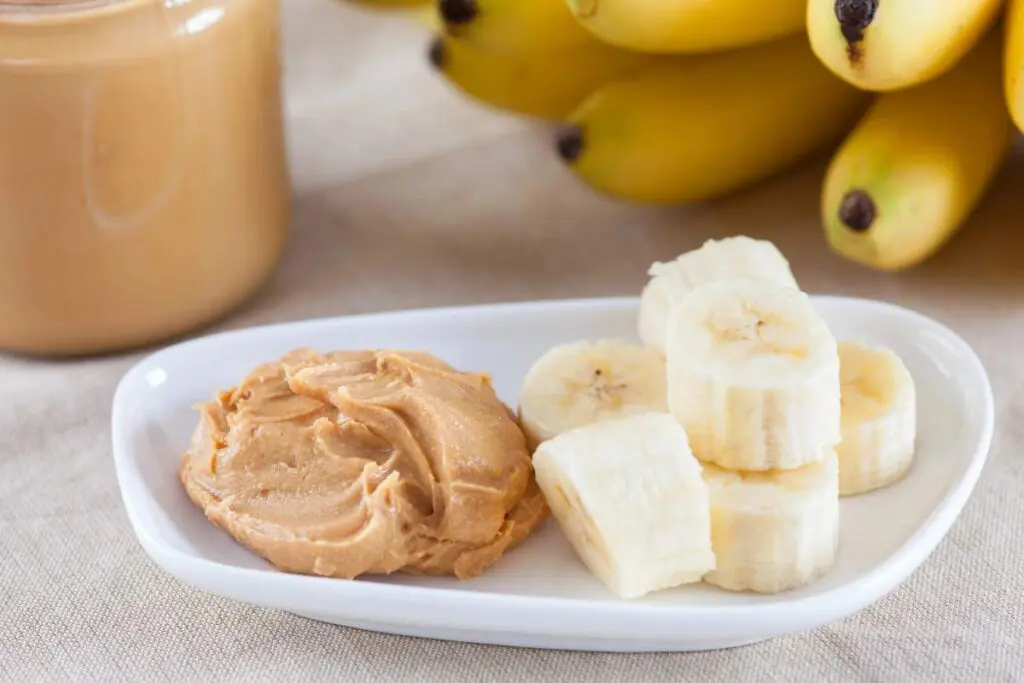 A plate with peanut butter and sliced bananas, with a jar of peanut butter and a bunch of bananas in the background.