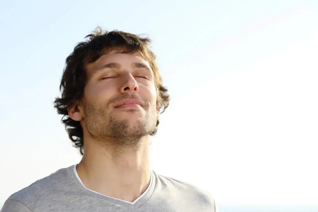 A man with short, curly hair is standing outdoors, eyes closed, with a serene expression on his face.