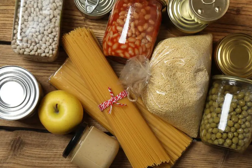 A variety of food items including pasta, jars of beans, grains, an apple, and cans on a wooden surface.