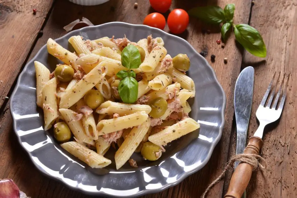A plate of pasta with penne, tuna, green olives, and fresh basil on a wooden table. Cherry tomatoes and cutlery are placed beside the plate.