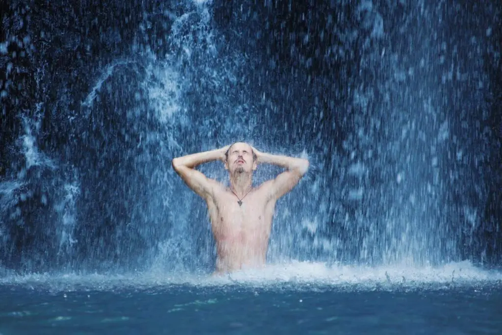 A person standing under a waterfall with water cascading around them, appearing serene and meditative.