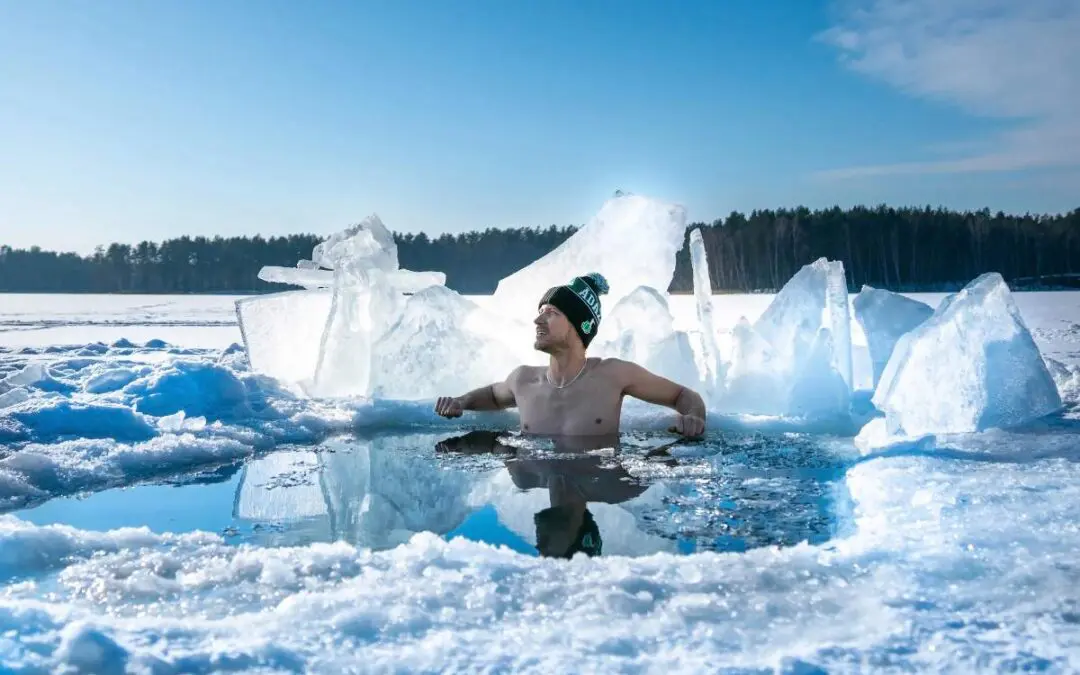 A shirtless man sitting in a thermal pool surrounded by ice and snow, wearing a beanie, with a blue sky and tree line in the background.