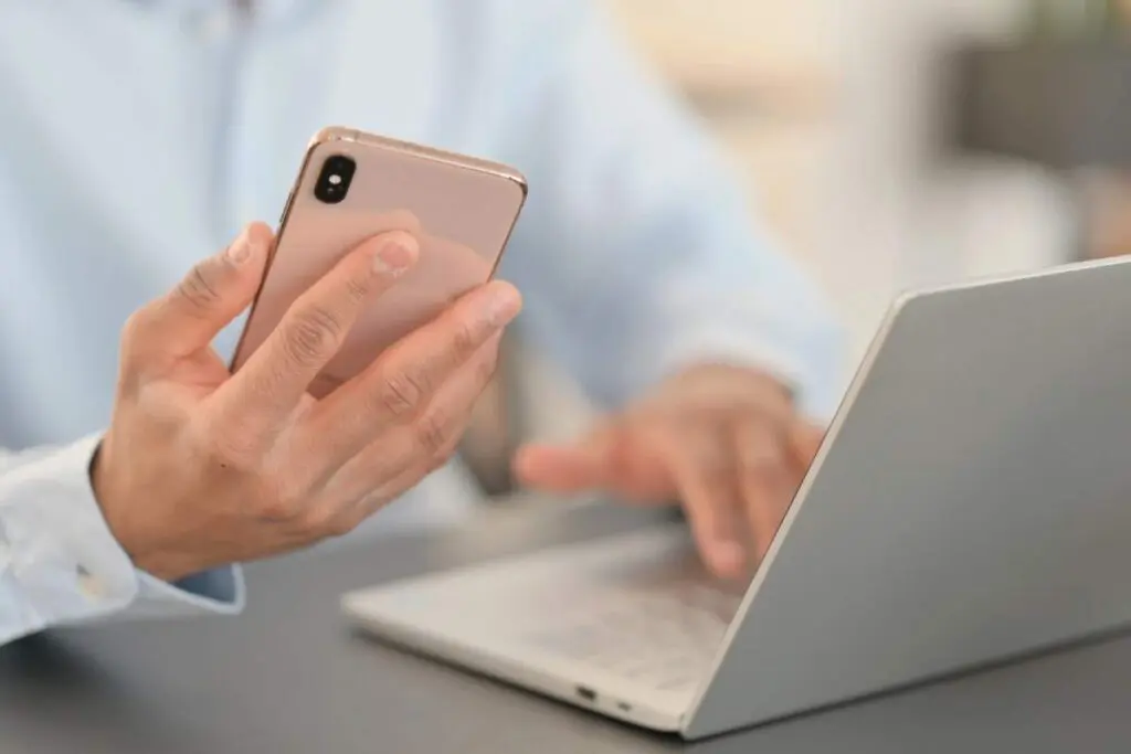 A person holding a smartphone in one hand while typing on a laptop with the other hand.
