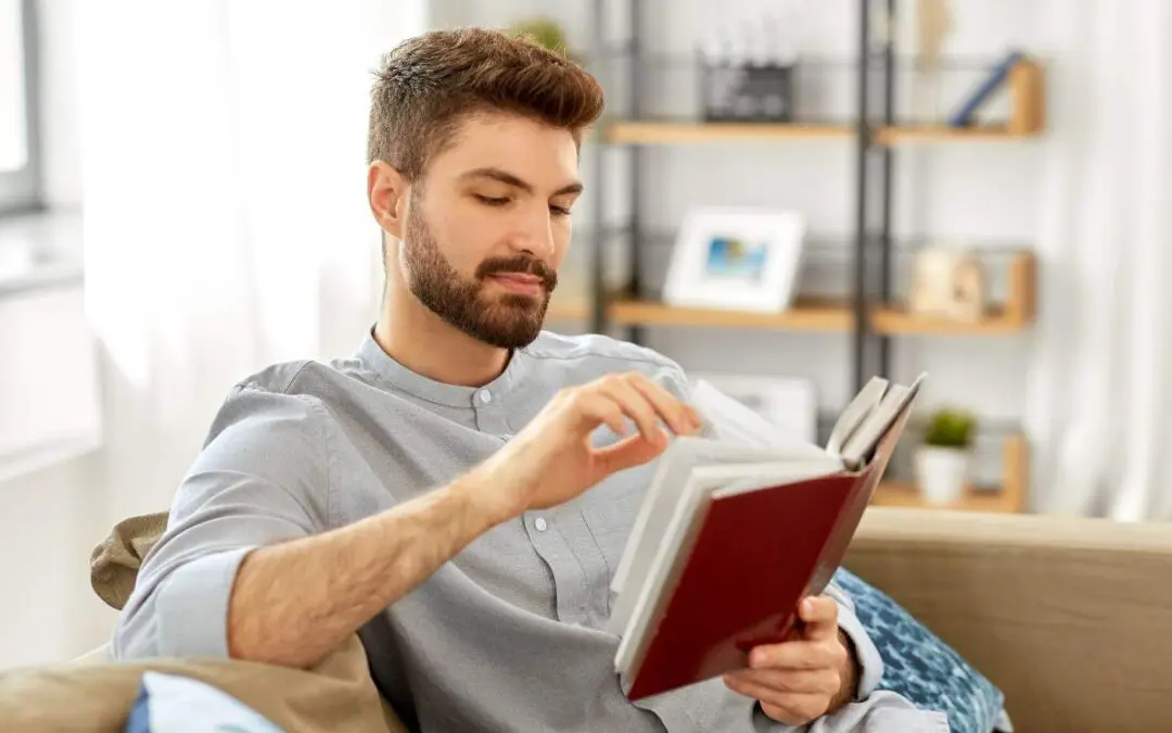 A young man with a beard is sitting on a couch and reading a book. He looks focused and engaged in his reading, with shelves visible in the background.