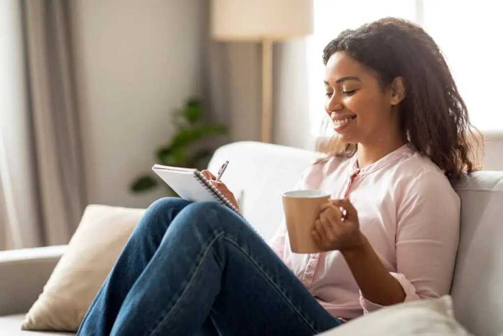 A smiling woman writing on a journal while holding a mug.