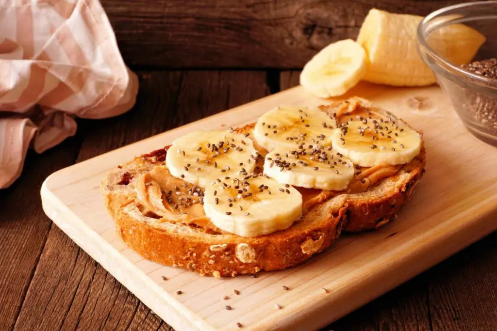 A slice of whole grain toast topped with peanut butter, banana slices, and chia seeds, placed on a wooden cutting board.