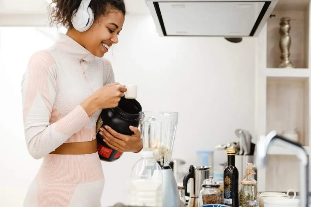 A woman wearing headphones prepares a protein shake in a kitchen, smiling as she holds a container of protein powder.