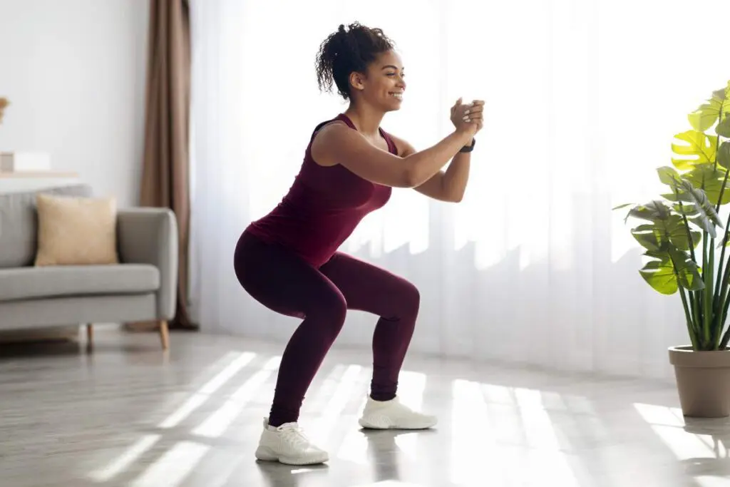 A woman in a maroon workout outfit performing a squat exercise indoors, with sunlight streaming through windows and a plant nearby.