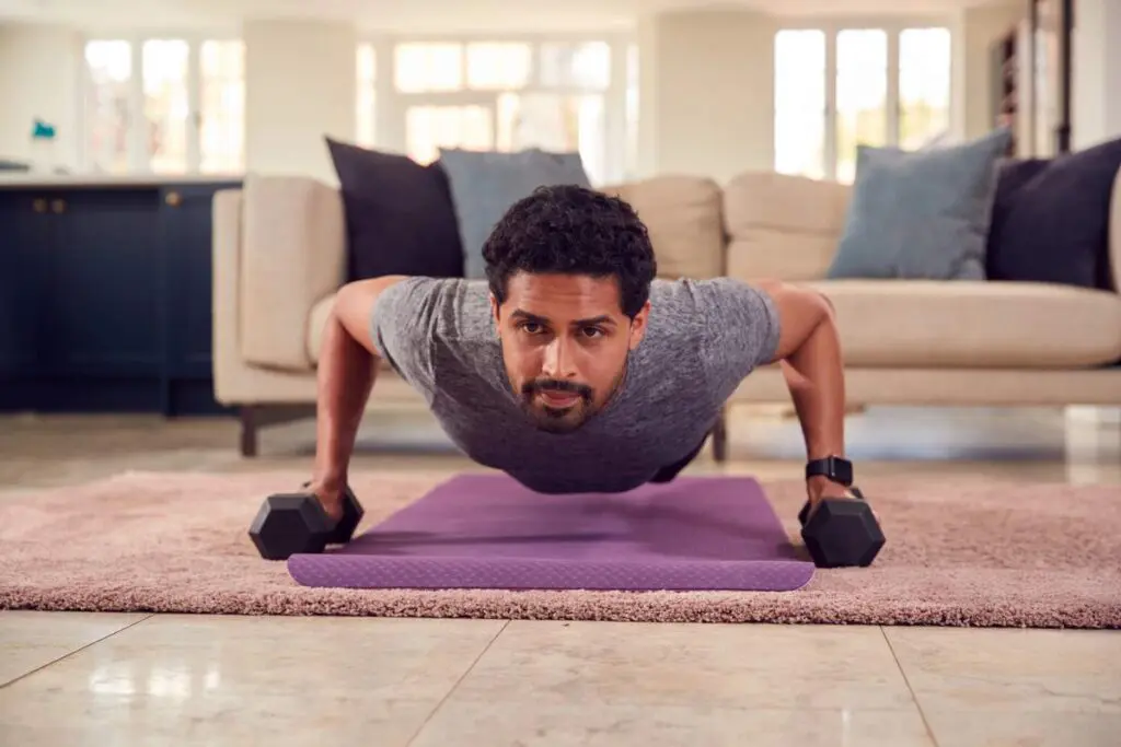 A man performing push-ups on a yoga mat in a living room, using dumbbells for support.