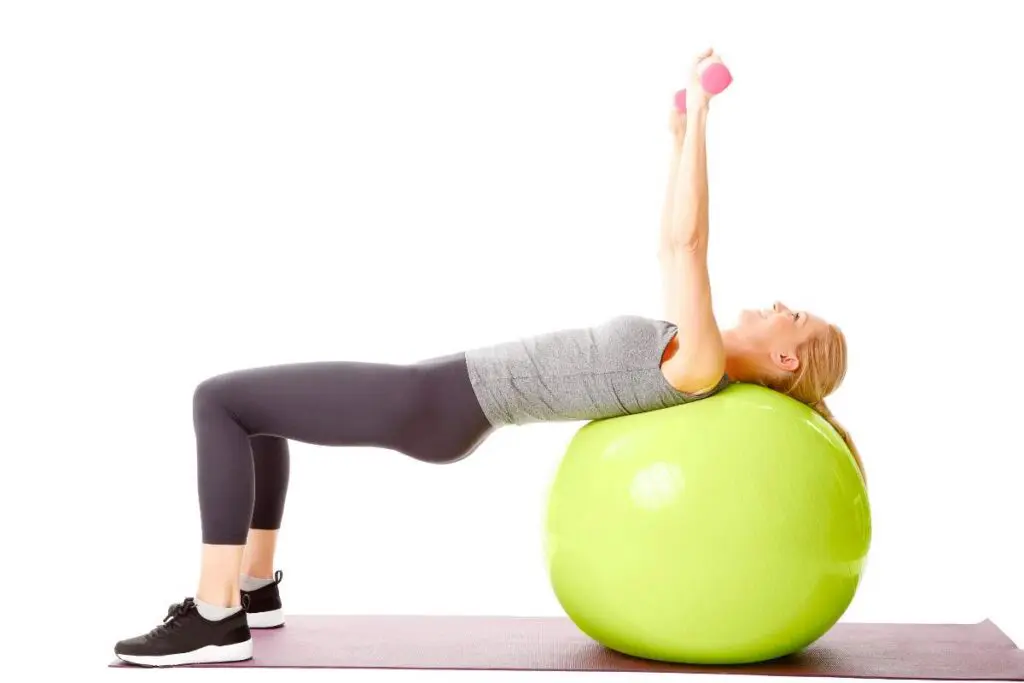 A woman performs an exercise on a green stability ball, holding pink dumbbells above her head while lying back.