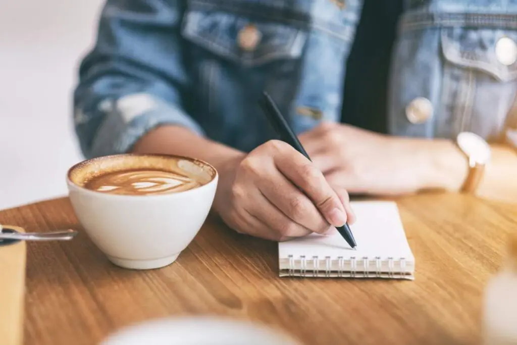 A person writing in a notepad with a cup of coffee on the table.