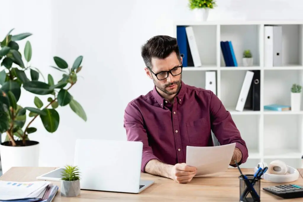 A man sitting at a desk, looking at a document. He is wearing glasses and a maroon shirt, with a laptop and office supplies nearby.