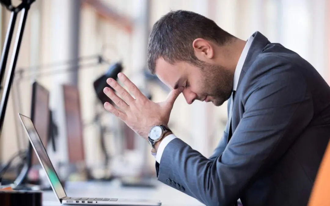 A man in a suit sitting at a desk with his hands on his forehead, looking stressed or overwhelmed, with a laptop open in front of him.