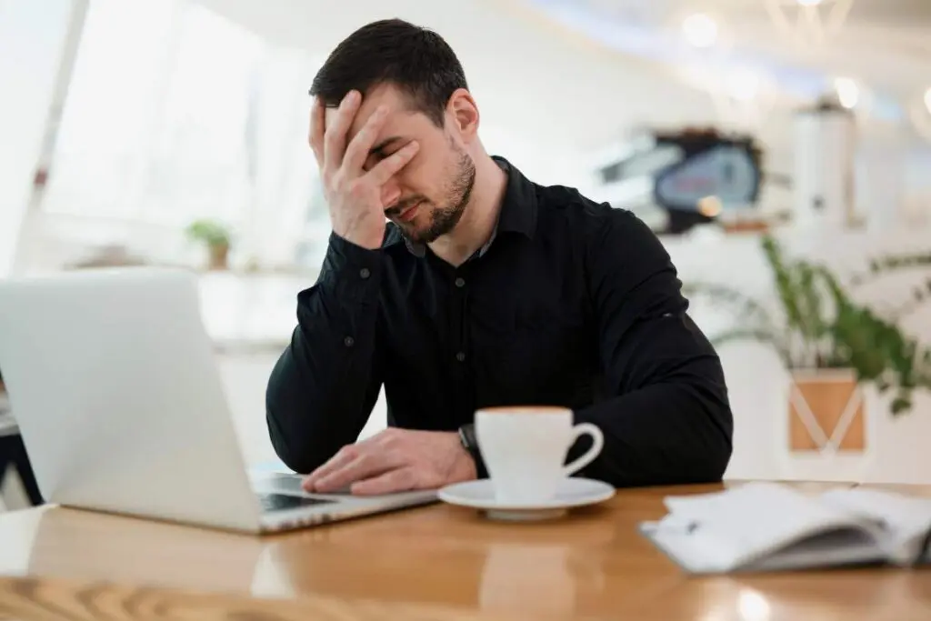 A man sitting at a table with a laptop, looking stressed and holding his forehead. There's a cup and saucer on the table beside him.