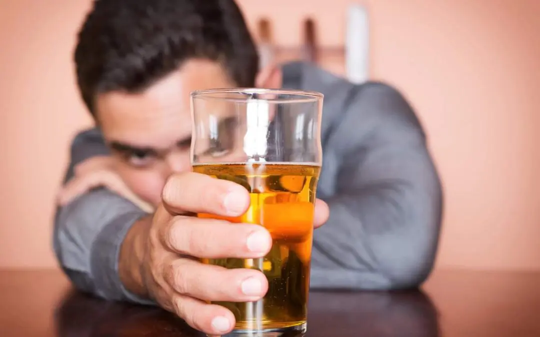 A man in a gray shirt is sitting at a table holding a glass of yellow beer, with his face partially obscured.