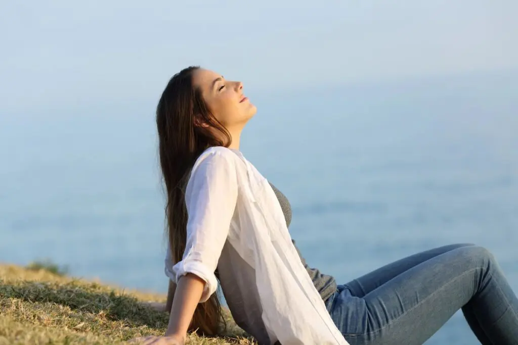 A woman sitting on the grass near a body of water, with her eyes closed and face turned up to the sun, appearing relaxed and at peace.