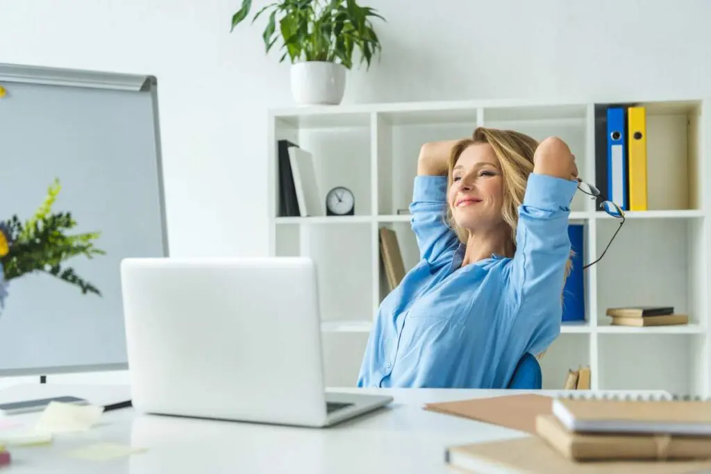 A woman sitting at a desk with a laptop, looking relaxed and smiling, with her hands behind her head.