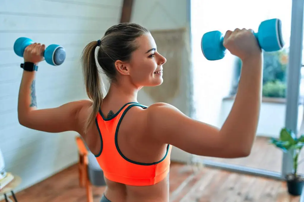 A woman in an orange sports bra is lifting light blue dumbbells in a bright, indoor setting.