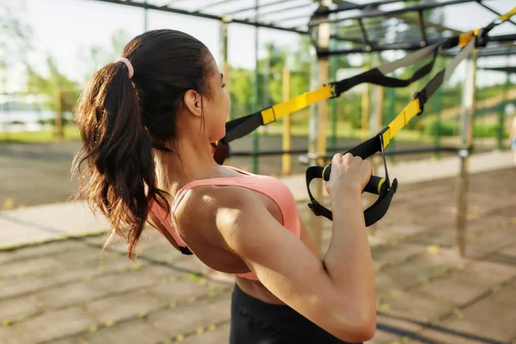 A woman performing a TRX exercise outdoors, pulling herself up using suspension straps.
