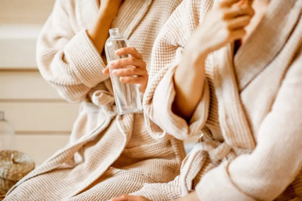 Two people wearing beige bathrobes, one holding a clear water bottle, sitting in a relaxing environment.