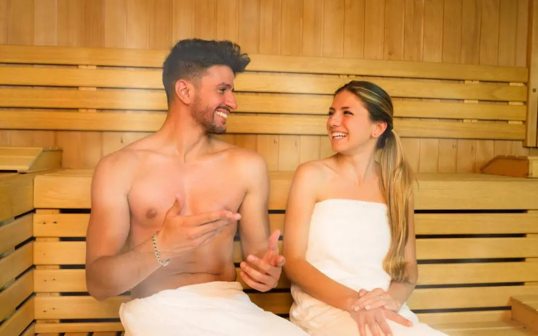 A man and a woman sitting in a sauna, both smiling and wearing towels.
