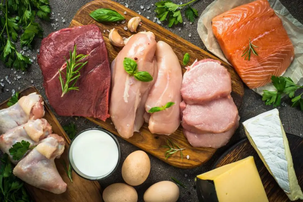 A variety of meats including beef, chicken, pork, and salmon arranged on a wooden board, accompanied by garlic, herbs, eggs, milk, and cheese.
