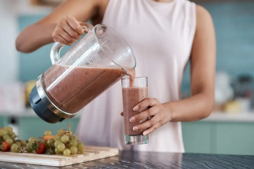 A person pouring a smoothie from a blender into a glass while standing in a kitchen.