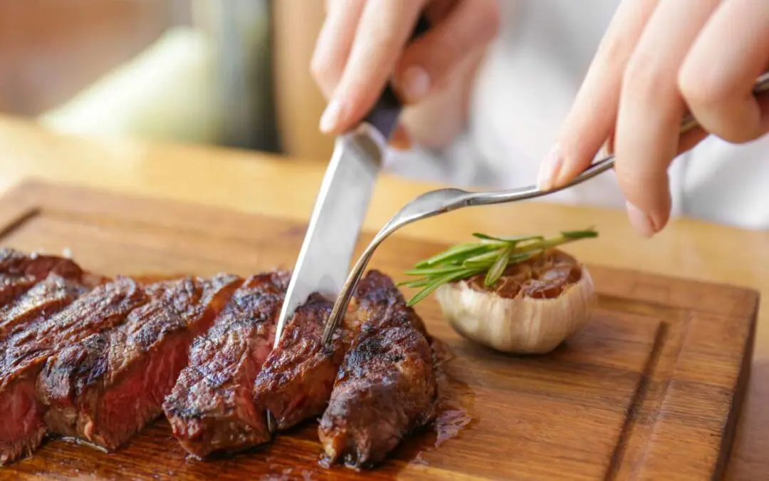 A person cutting into a piece of steak on a wooden cutting board, accompanied by roasted garlic and garnished with chives.