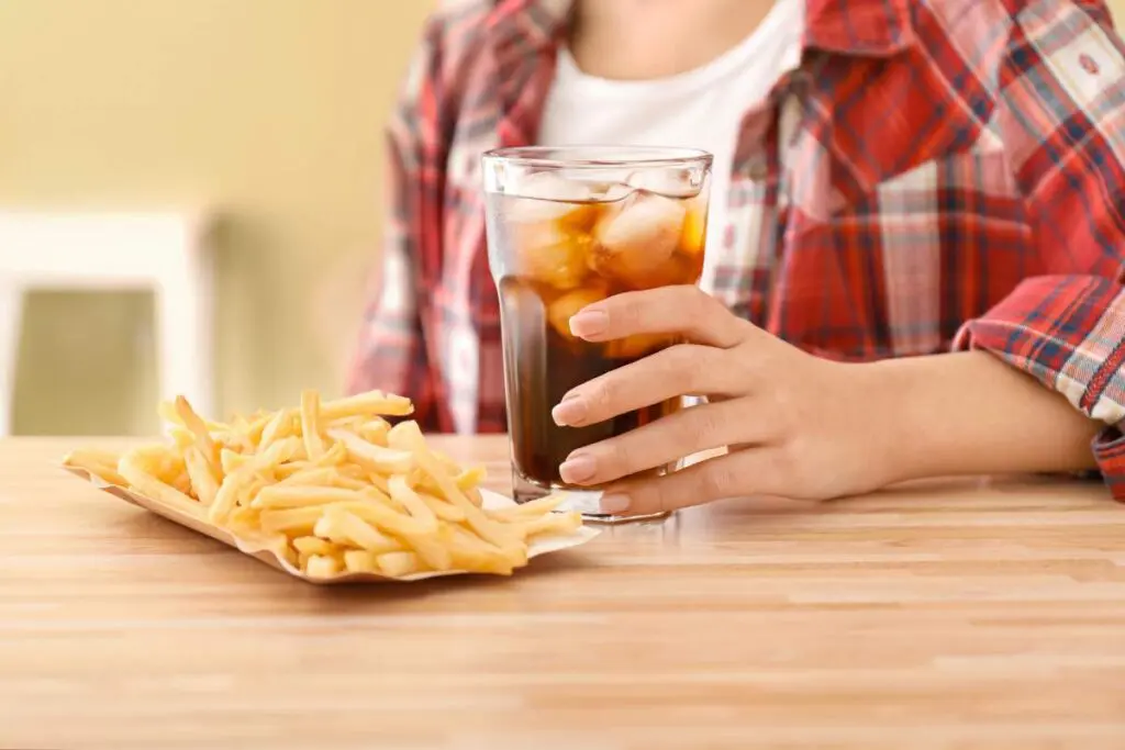 A person wearing a plaid shirt holding a glass of iced drink with a plate of French fries on a wooden table.