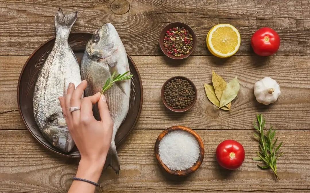 A hand preparing two whole fish on a wooden table, surrounded by various ingredients including spices, garlic, lemon slices, tomatoes, and herbs.