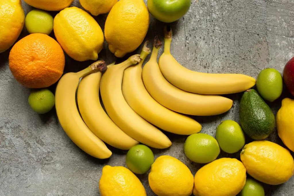 A variety of fruits on a textured surface, including bananas, lemons, limes, green apples, an orange, and an avocado.