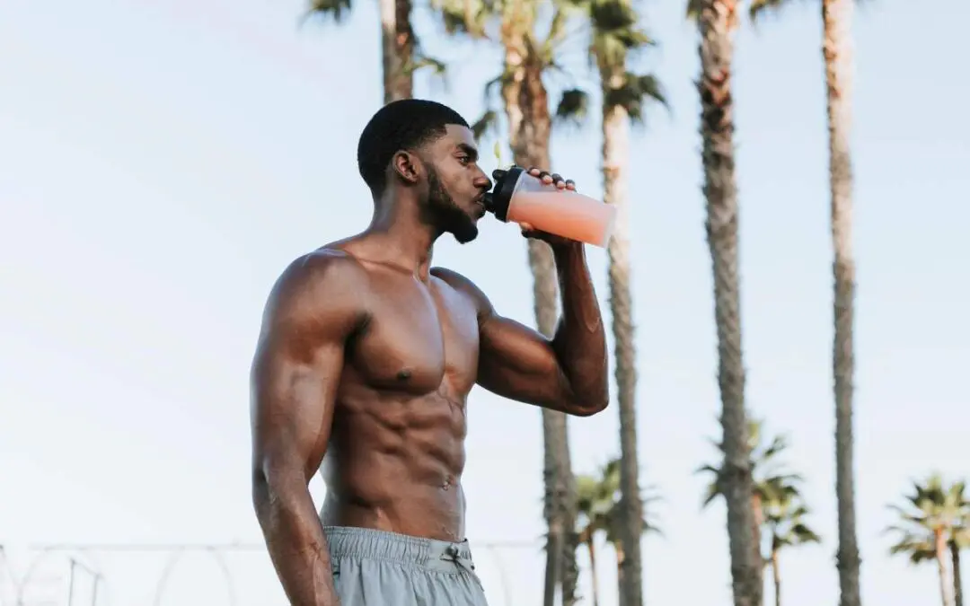 A muscular man with an athletic build drinks from a shaker cup outdoors, surrounded by palm trees.