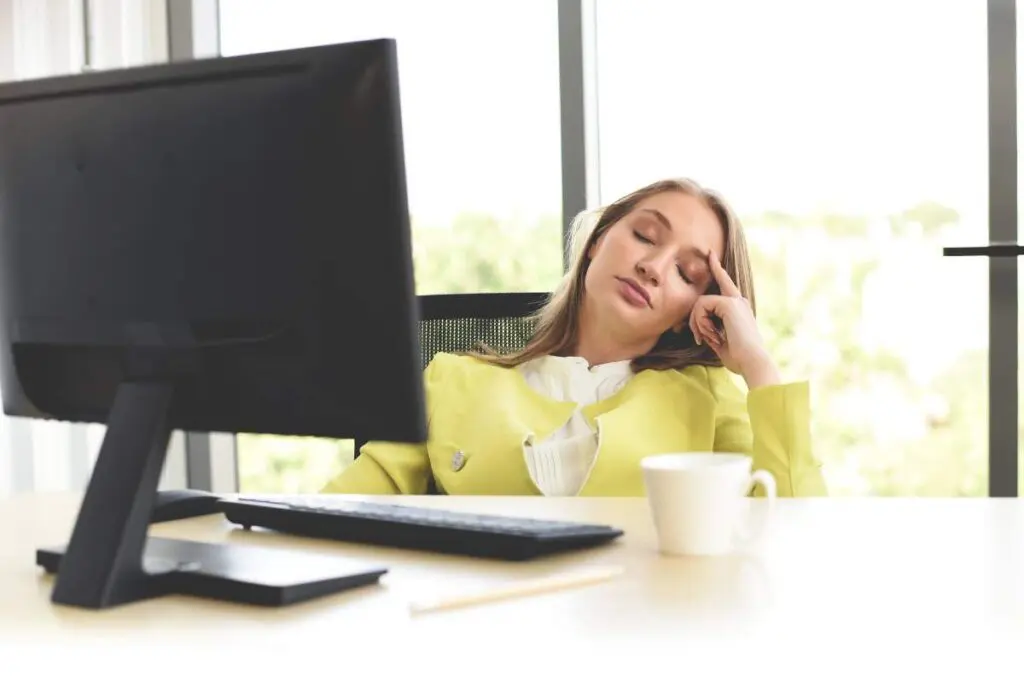 A woman in a bright yellow blazer rests her head against her hand while seated at a desk with a computer monitor, appearing tired or distracted.