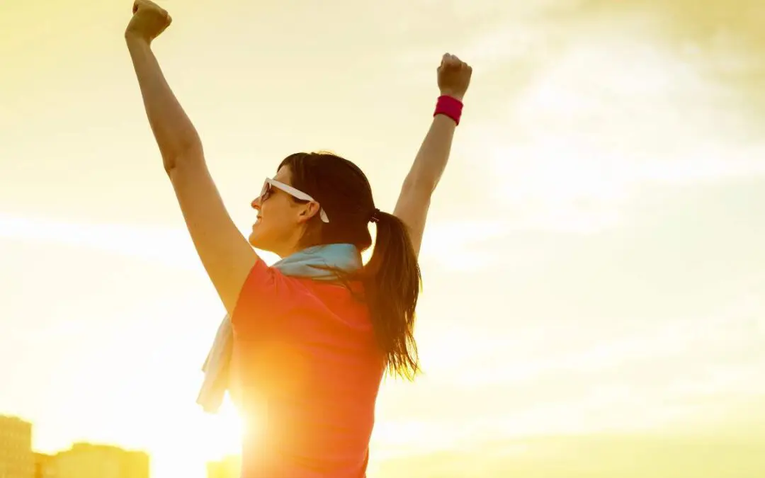 A woman raises her arms in celebration against a sunset background.