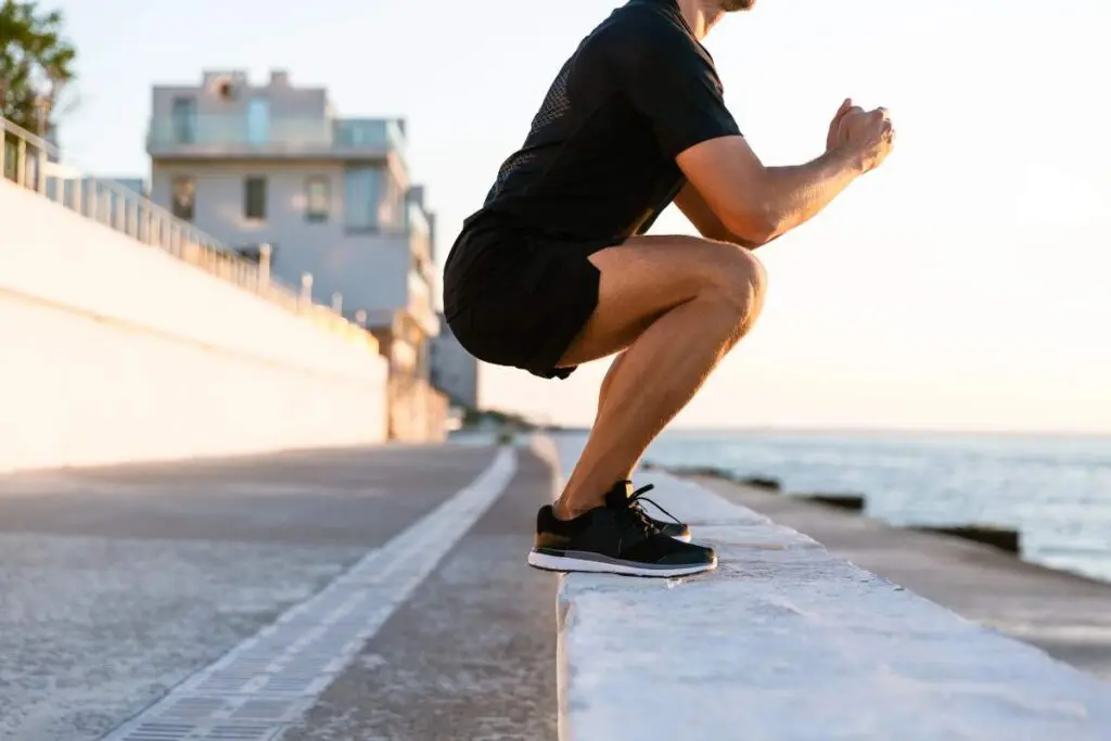 A man performing a squat exercise on a ledge near a body of water during sunset.