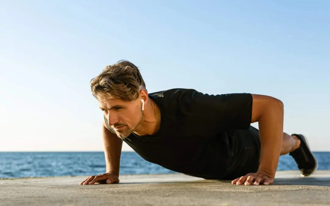 A man performing push-ups outdoors by the water, wearing earbuds.