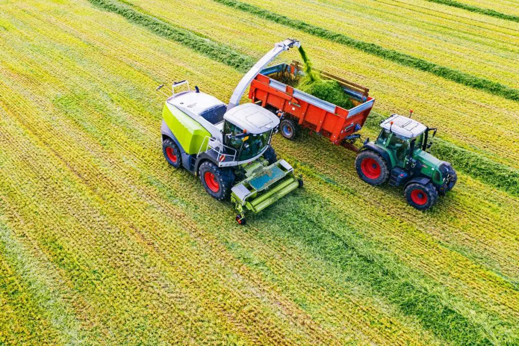 Aerial view of a green harvesting machine and a tractor with a trailer in a field, working on freshly cut grass.