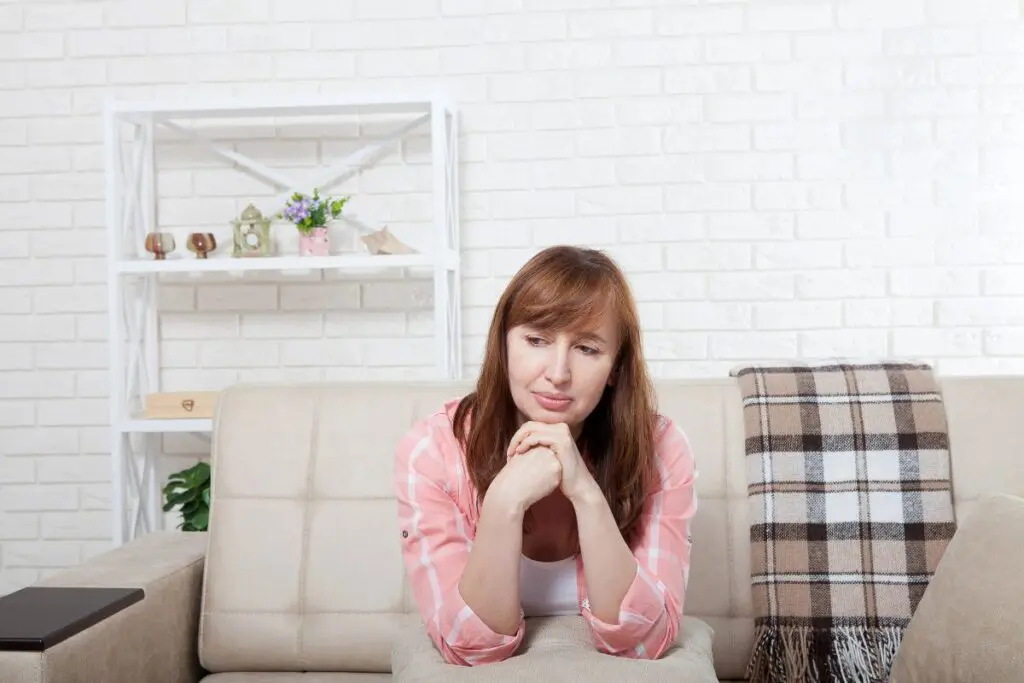A woman sitting on a couch with a thoughtful expression, resting her chin on her hands, in a cozy living room setting.
