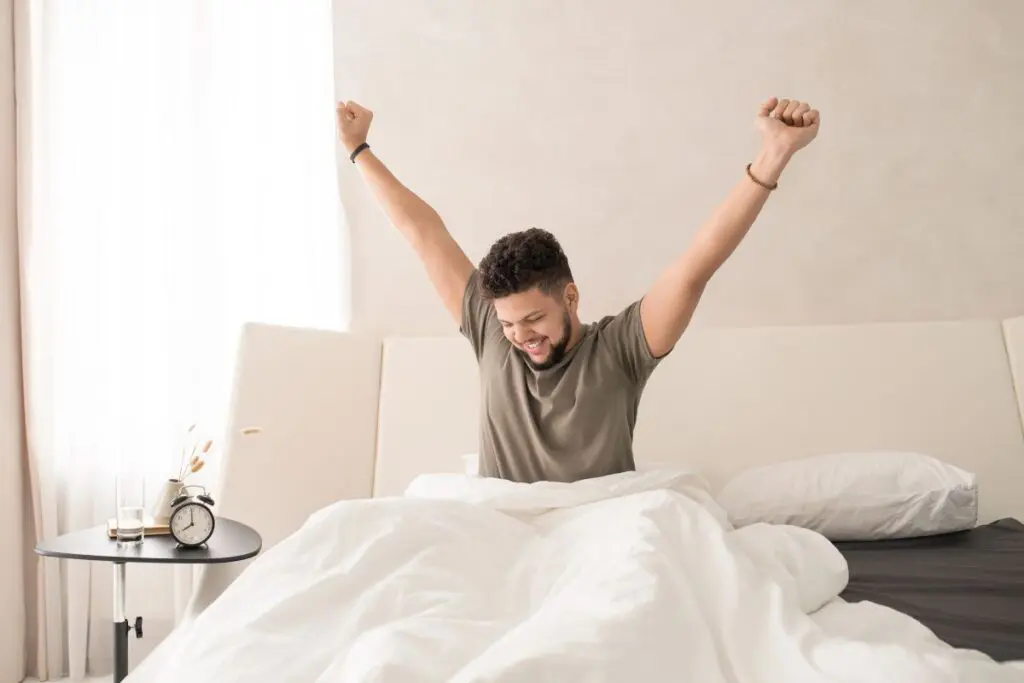 A man stretching in bed with a smile, morning light coming through the window.