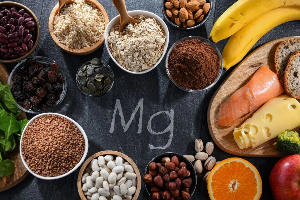 A variety of foods rich in magnesium, including nuts, seeds, grains, fruits, and vegetables, arranged on a dark surface with "Mg" written in the center.