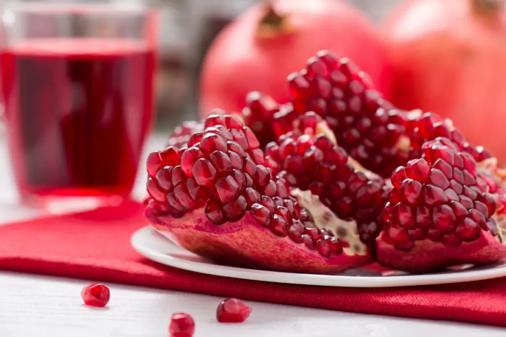 A plate of pomegranate seeds with a glass of pomegranate juice in the background, on a red cloth.