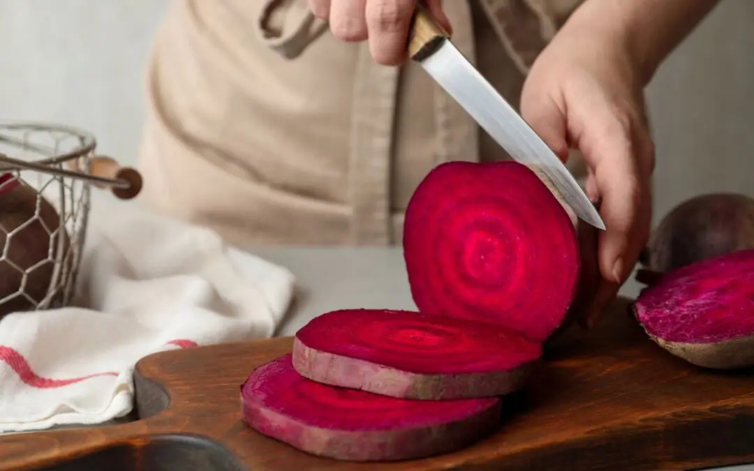 A person's hand slicing a bright red beet on a wooden cutting board, with additional beets and a basket in the background.